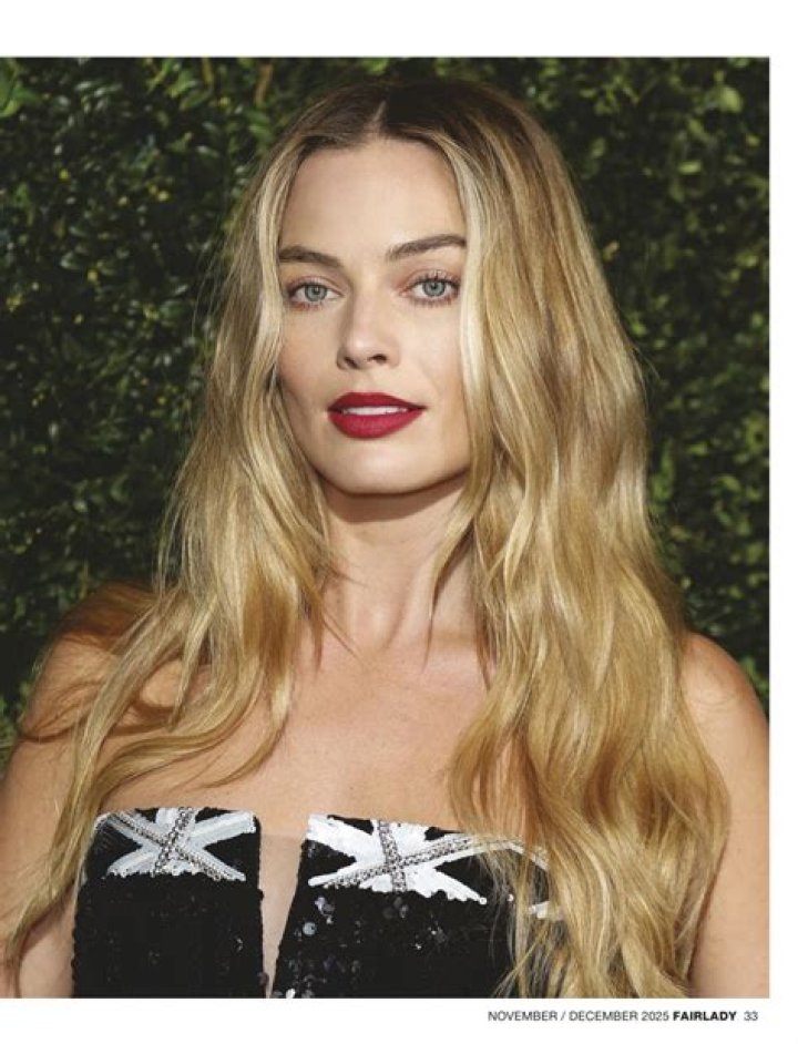 Margot Robbie is wearing black and sitting on a couch, while speaking to someone across from her. 
